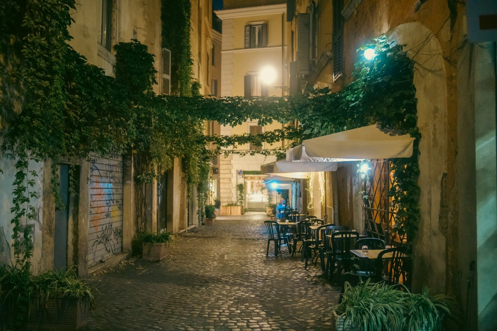 Evening view of a charming, narrow cobblestone street in Trastevere, with ivy-clad ochre buildings, outdoor tables, and soft street lighting.