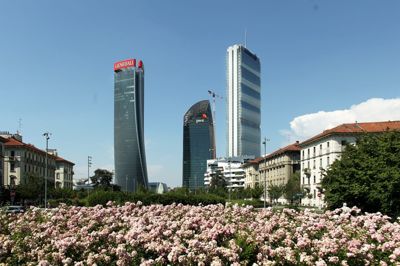 I grattacieli Tre Torri nel quartiere CityLife di Milano si innalzano dietro un campo fiorito di fiori rosa, incorniciati da edifici tradizionali della città sotto un cielo sereno.