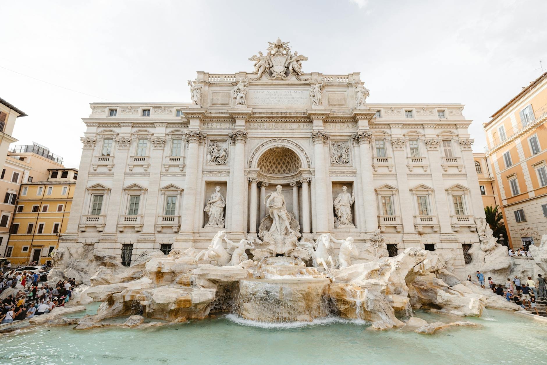 Wide, front-facing view of the entire Trevi Fountain and surrounding buildings under daylight, with turquoise water and some visitors visible at the edges.