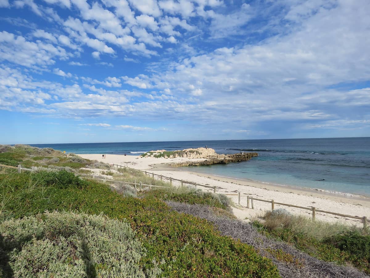 Wide landscape view of Trigg Beach with limestone outcrop, white sand, Indian Ocean waves, low dunes, and blue sky with scattered clouds in Perth, Australia.