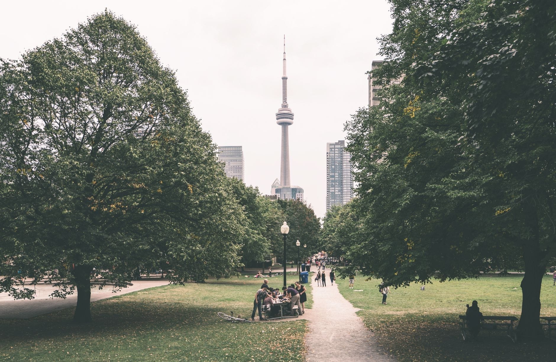 ทางเดินกว้างที่มีต้นไม้ใหญ่เขียวสองข้าง คนกำลังนั่งบนม้านั่ง เห็น CN Tower และเส้นขอบฟ้าโตรอนโตในวันที่เมฆมาก