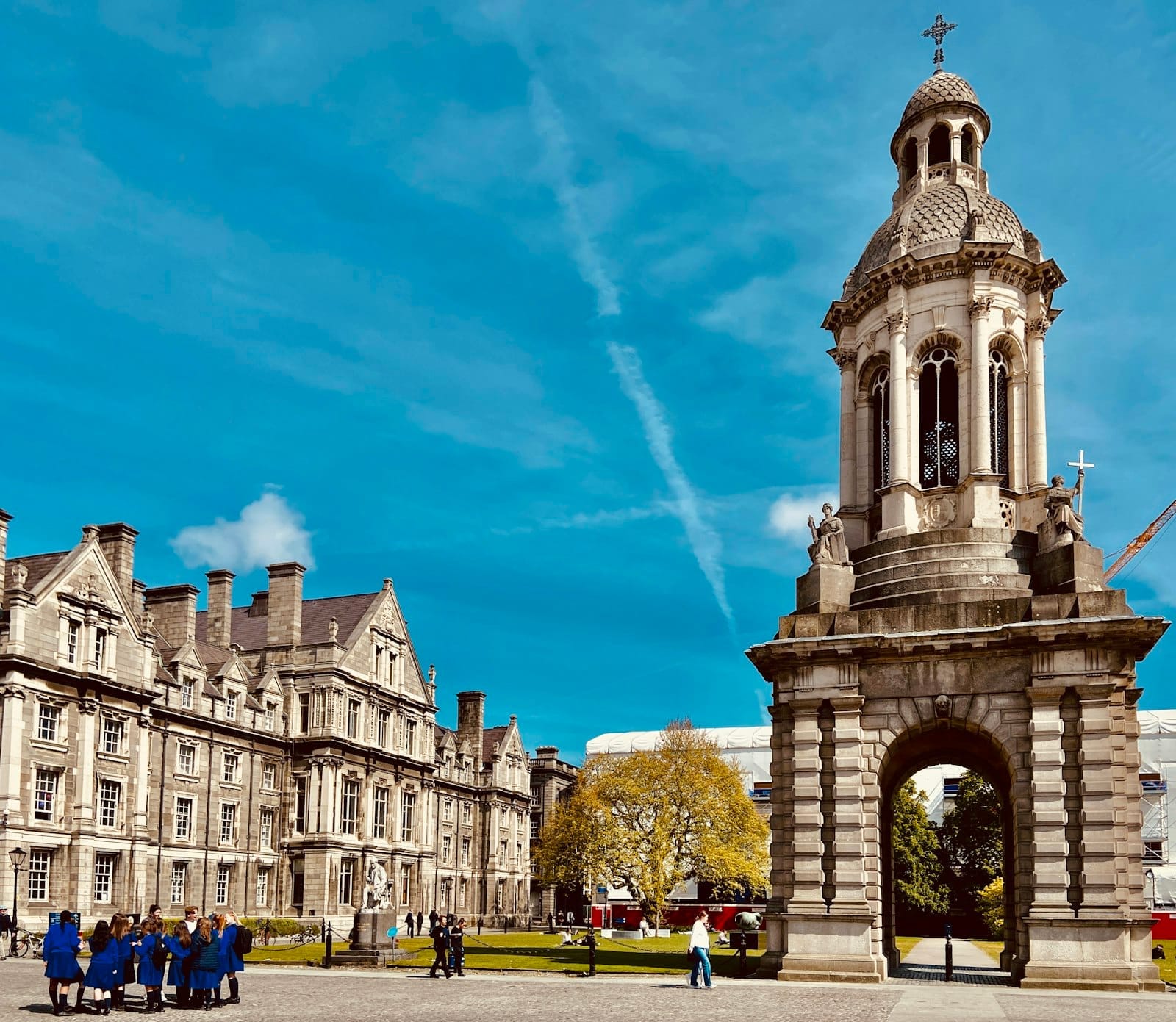 ลานของ Trinity College Dublin กับหอระฆังสัญลักษณ์ ตึกหินเก่าแก่ ท้องฟ้าสีฟ้า และกลุ่มผู้คนเดินชมบรรยากาศ