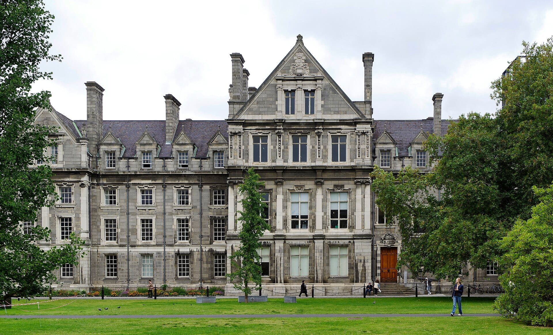 Storica facciata in pietra del Trinity College Dublin con architettura georgiana, lussureggiante prato verde e visitatori che passeggiano in una giornata nuvolosa.