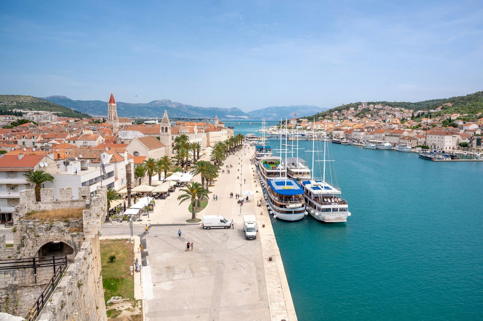 วิวริมน้ำประวัติศาสตร์ของ Trogir พร้อมอาคารหินเรียงราย ทางเดินเลียบชายหาดที่ร่มรื่นด้วยต้นปาล์ม เรือจอดเทียบท่า และน้ำสีฟ้าใสใต้ท้องฟ้าที่แจ่มใส