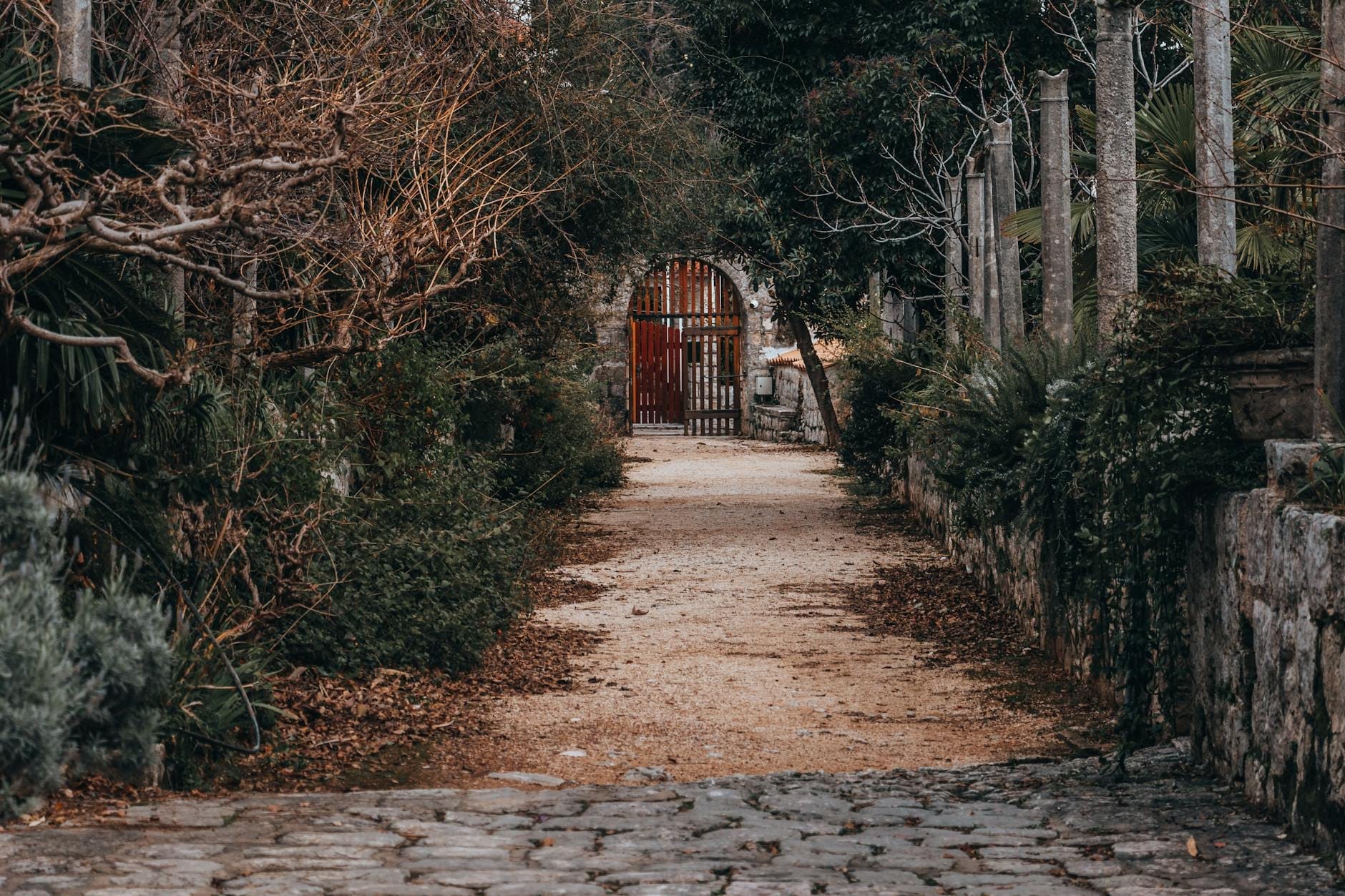 Sendero de piedra entre exuberante vegetación y columnas que lleva a una arcada de puerta roja en el Arboreto de Trsteno, con todo el encanto de un jardín de otro tiempo cerca de Dubrovnik.