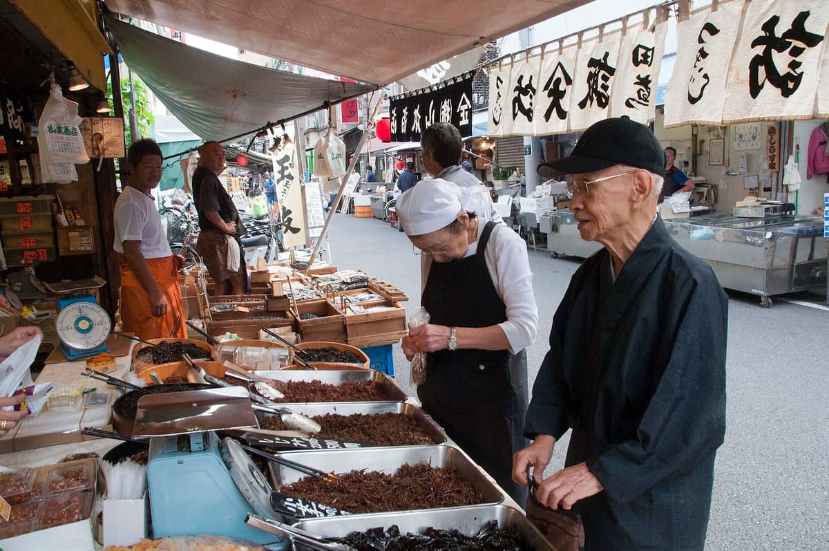 Street-side market stalls at Tsukiji Outer Market with various foods and shoppers, showcasing a lively atmosphere and classic handwritten banners.