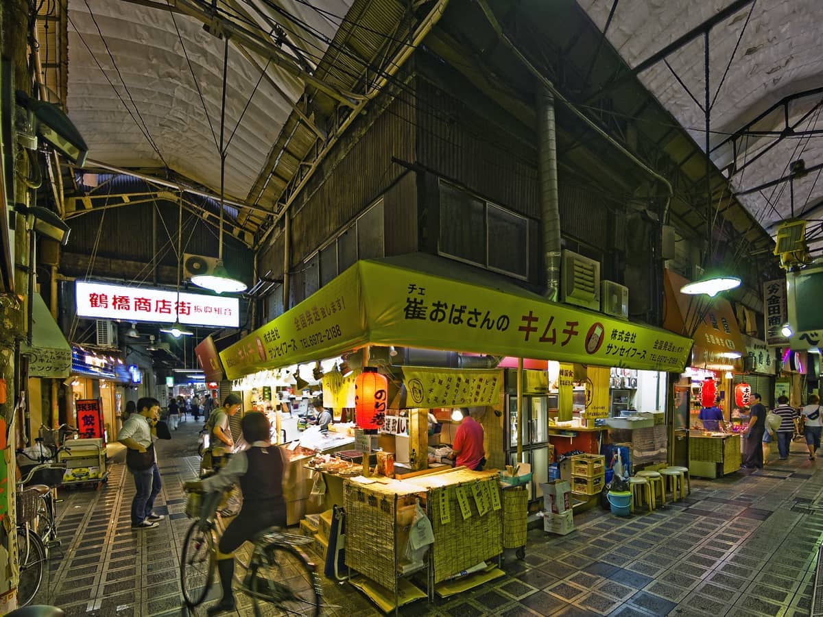 Covered street in Tsuruhashi Korea Town at night, with yellow-lit food stalls, traditional signs, and people shopping along the walkway.