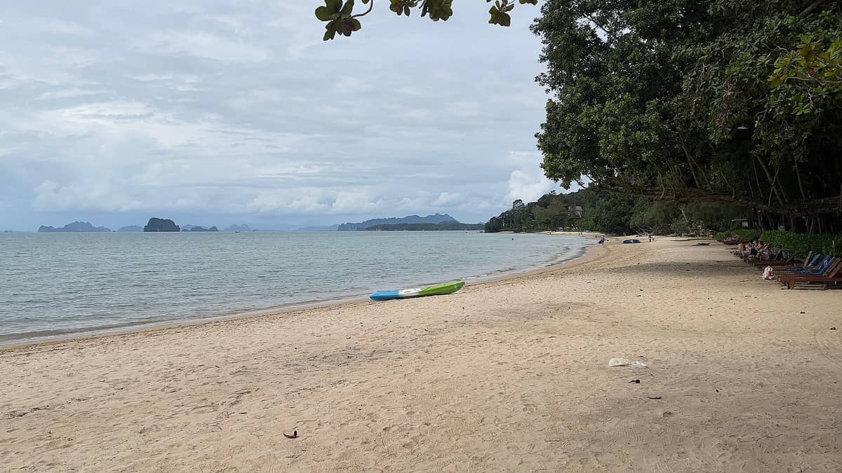 La amplia y arenosa playa Tubkaek con aguas calmas, algunas tumbonas dispersas, árboles inclinados y lejanas islas de piedra caliza kárstica bajo un cielo nublado en Krabi, Tailandia.