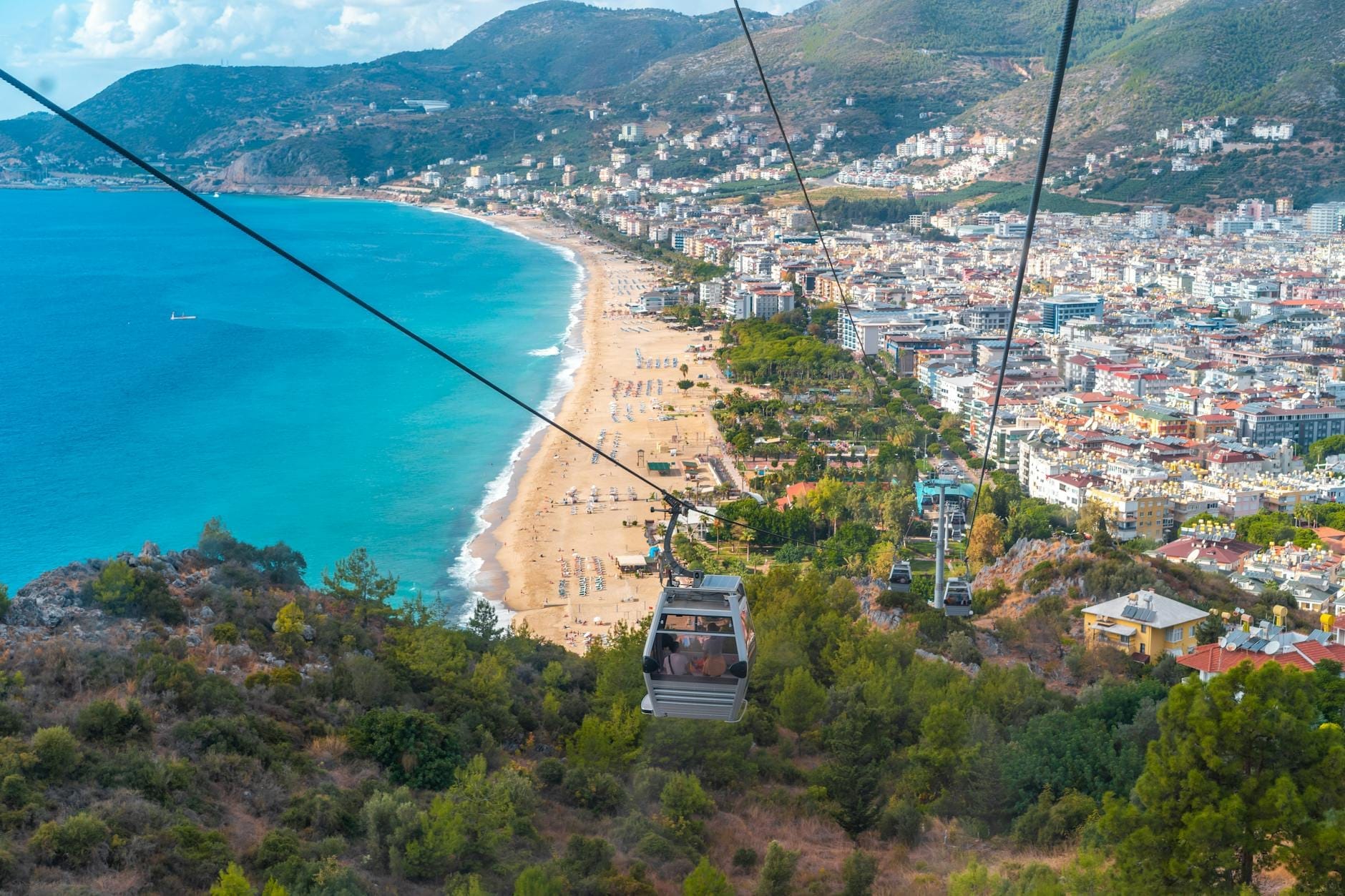 Una cabinovia scende su colline verdeggianti con una vista panoramica sulla spiaggia di Konyaaltı ad Antalya, il profilo urbano e i lontani monti Tauro sotto un cielo azzurro.