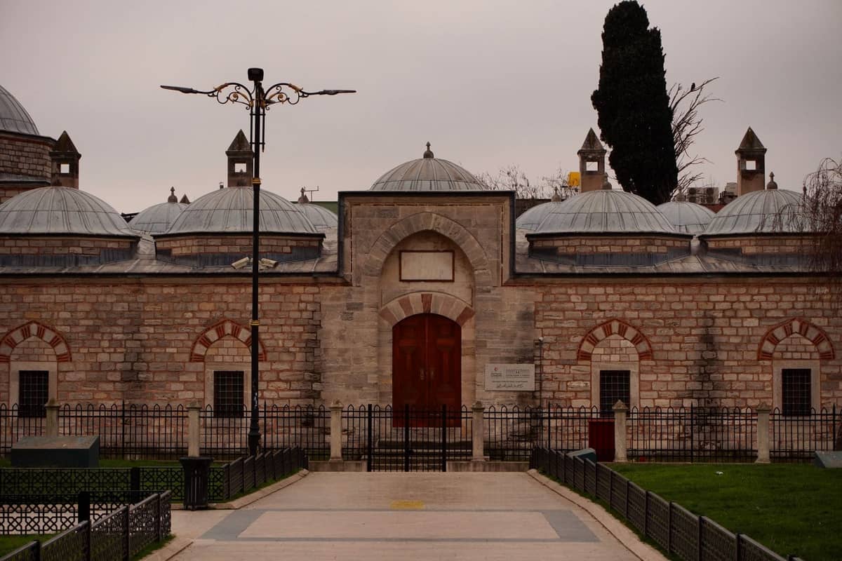 Entrada principal de un histórico hammam de Estambul con cúpulas de piedra y una puerta de madera arqueada, vista desde la calle.