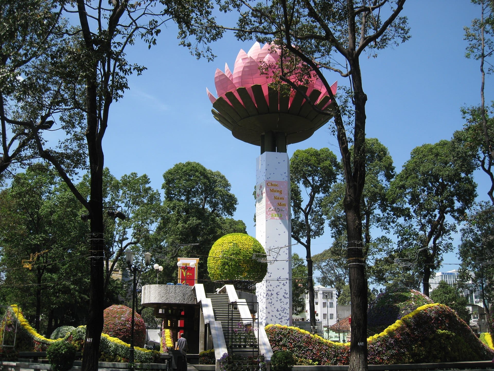 La Rotonda del Lago de la Tortuga presenta una alta columna de concreto con una escultura de loto rosa, rodeada de exuberantes árboles y coloridas exhibiciones florales bajo un cielo azul.