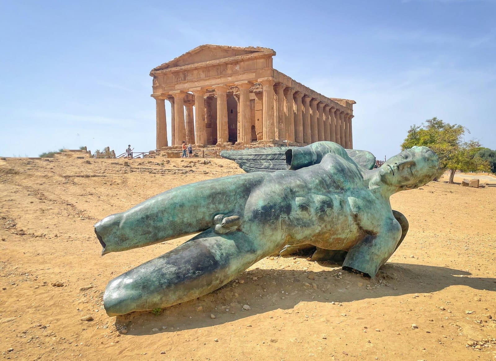Antiker griechischer Tempel in Agrigento mit einer großen gefallenen Bronzestatue im Vordergrund unter einem klaren Himmel.