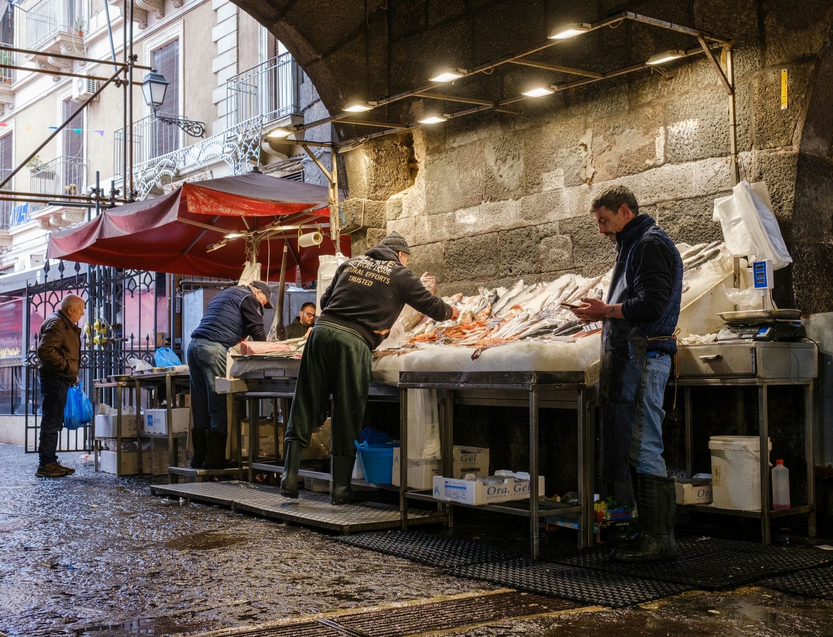 Händler und Käufer auf einem Fischmarkt im Freien in Catania mit frischen Meeresfrüchten, die auf Ständen in einer historischen Steinumgebung ausgestellt sind.