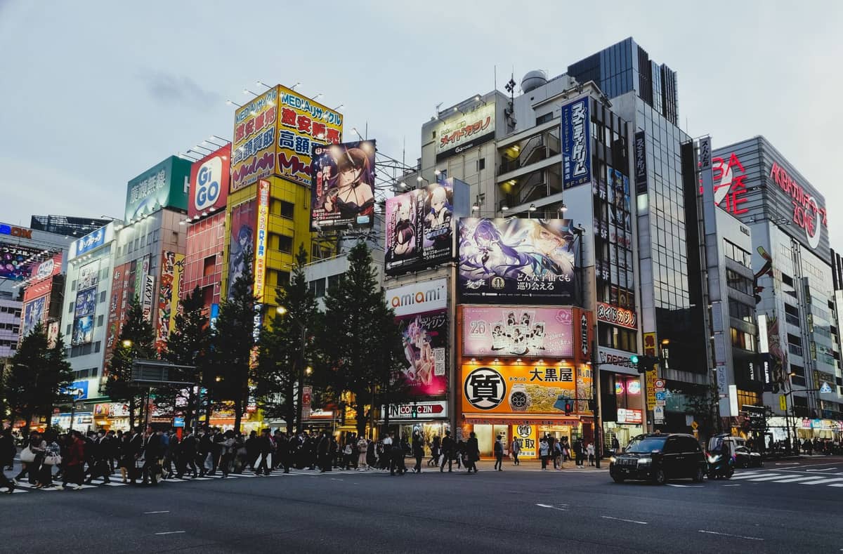 Akihabara intersection at dusk, crowded with people crossing streets and surrounded by colorful anime billboards and tall buildings. A lively Tokyo city scene.