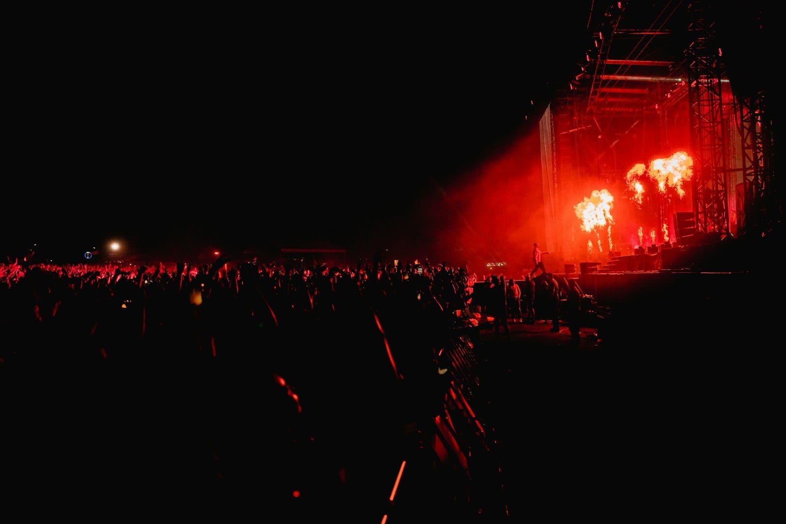 Grande multidão em um festival de música noturno com luzes vermelhas intensas no palco e pirotecnia, transmitindo a energia e a animação de um evento ao ar livre.