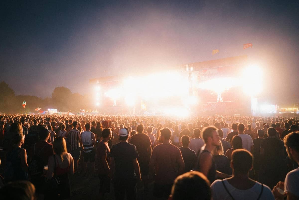 Large outdoor music festival crowd at night facing a brightly lit stage, creating a vibrant night-time concert atmosphere.
