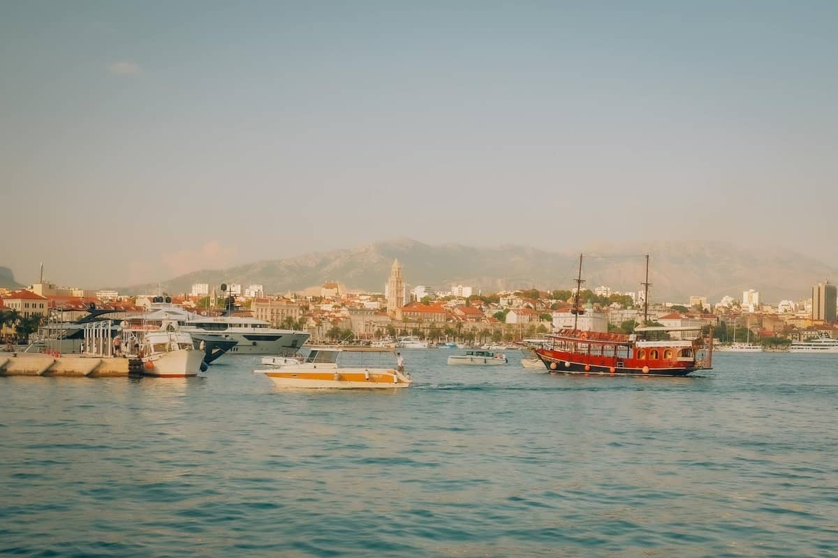 Several boats and yachts in Split's harbor with the recognizable old town skyline and mountains in the distance under clear skies.