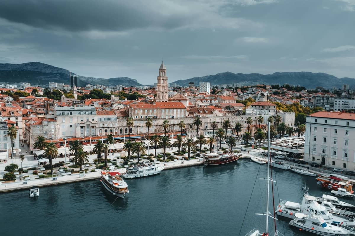 A wide view of Split’s historic waterfront featuring Diocletian’s Palace, old town, palm trees, and boats in the harbor under a dramatic cloudy sky.