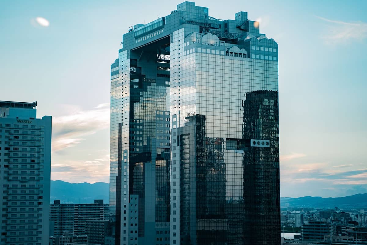 A striking landscape view of the Umeda Sky Building in Osaka, showing its twin towers connected at the top with the distinctive circular observatory against a blue sky.