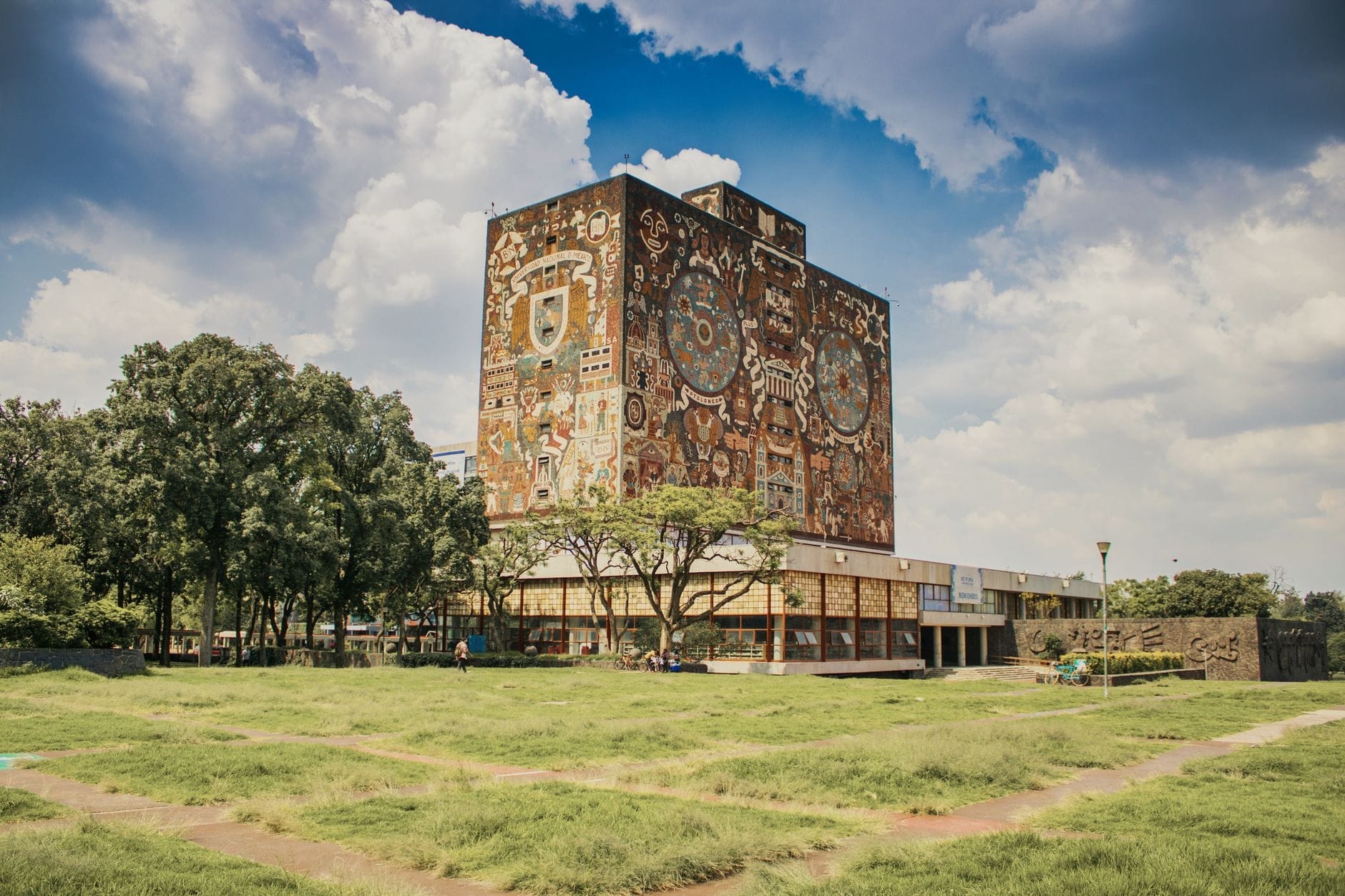 Vista panoramica della Biblioteca Centrale della UNAM con il suo iconico murales, circondata da prati verdi e alberi sotto un cielo parzialmente nuvoloso nel pomeriggio.