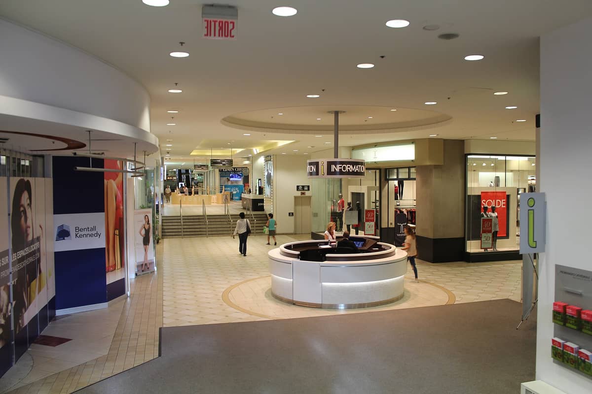 Weitwinklige Aufnahme eines Informationsschalters und einer Einkaufspassage in Montreals Underground City mit Geschäften, Beschilderung und Fußgängern.
