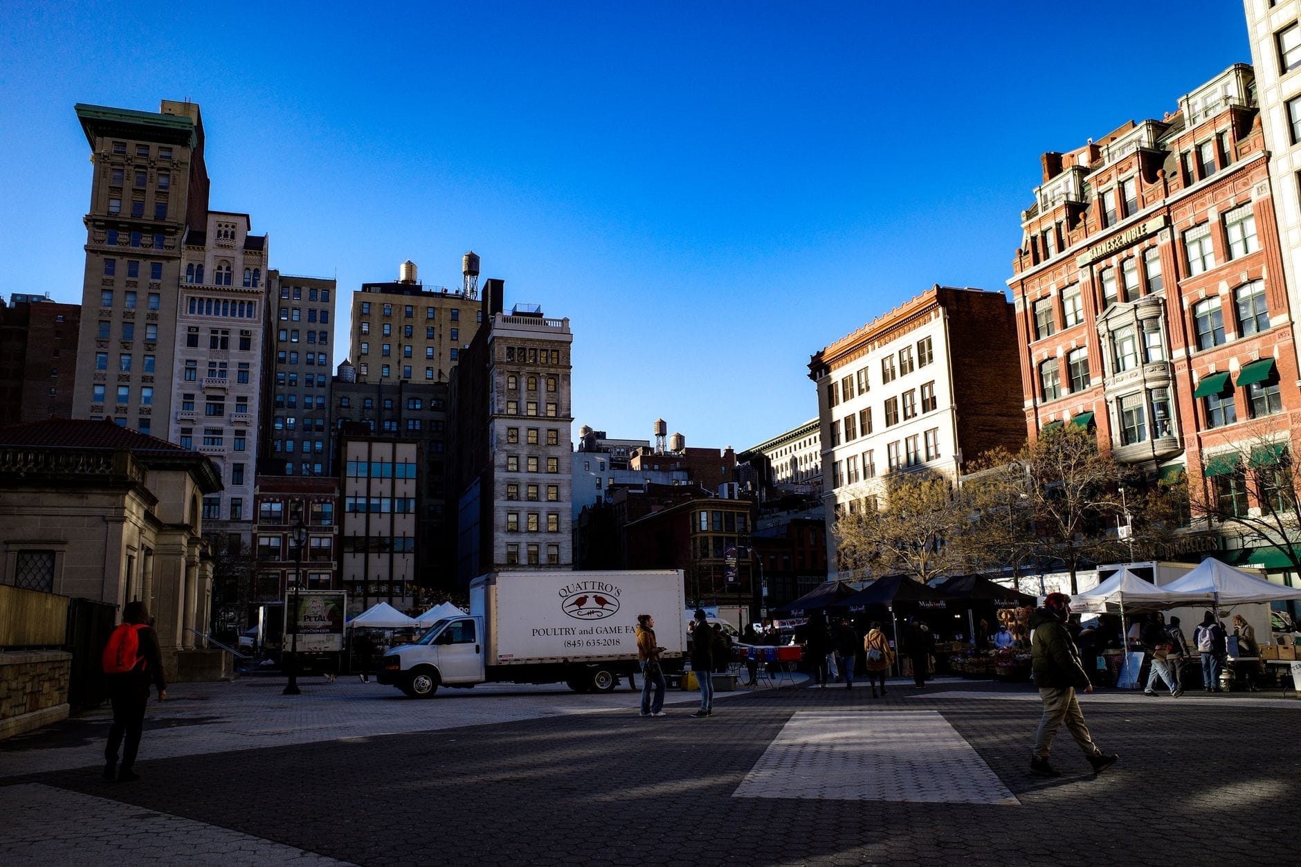 Il Greenmarket di Union Square con le tende bianche dei venditori, un furgone del mercato e persone che curiosano in una piazza aperta circondata dagli edifici di Manhattan sotto un cielo azzurro.