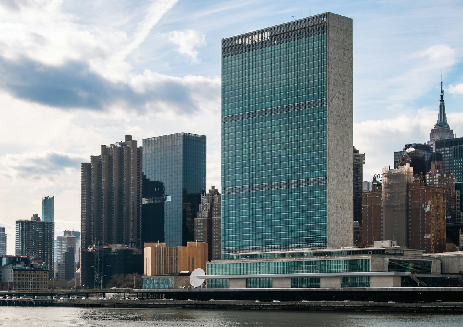 Vista della sede delle Nazioni Unite dall'altra sponda dell'East River, con il palazzo principale dell'ONU e lo skyline di Midtown Manhattan sotto un cielo parzialmente nuvoloso.