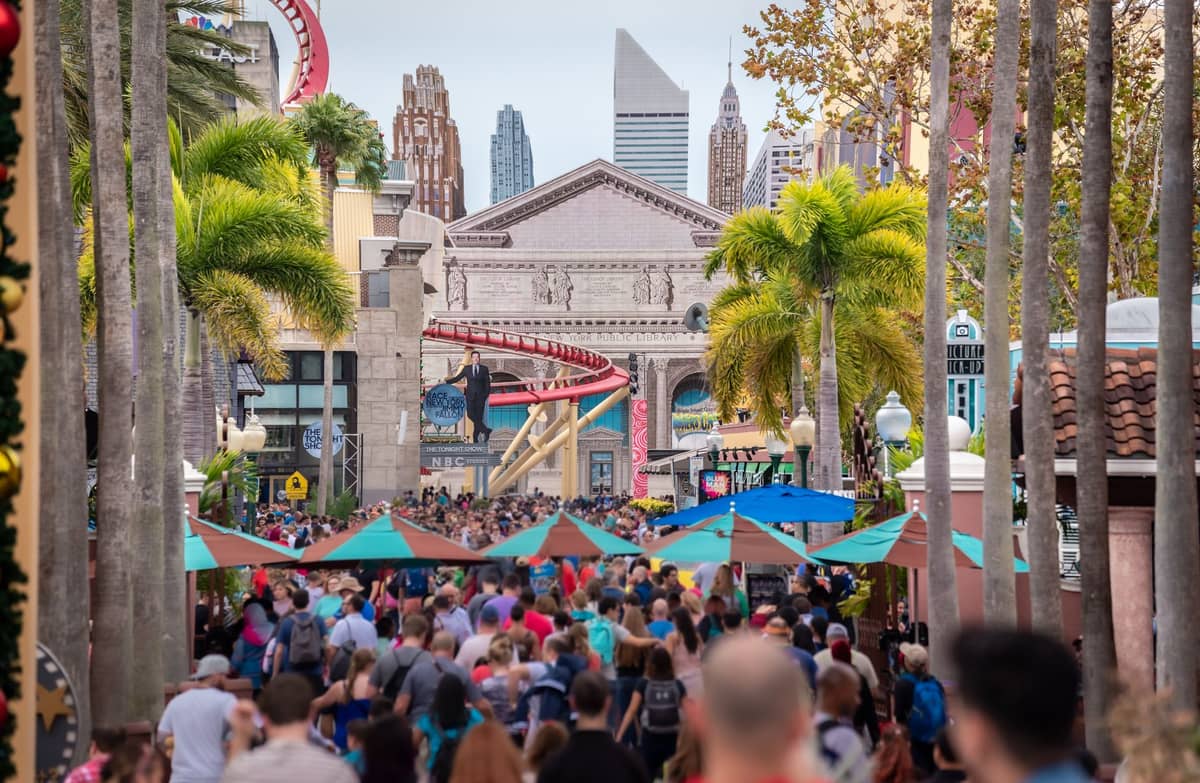 A bustling crowd walks down the main thoroughfare of Universal Studios Florida, with palm trees, themed buildings, and roller coaster tracks in the background.