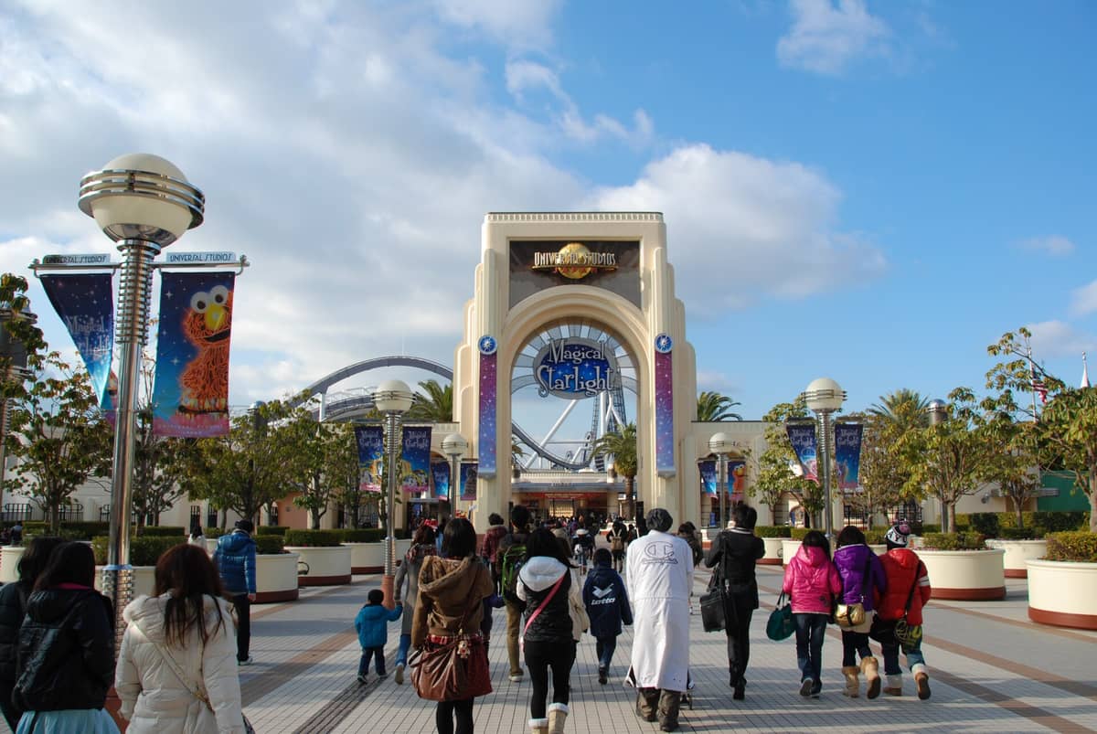 Entrance gate of Universal Studios Japan with crowds of visitors and themed banners, roller coaster tracks visible in the background.