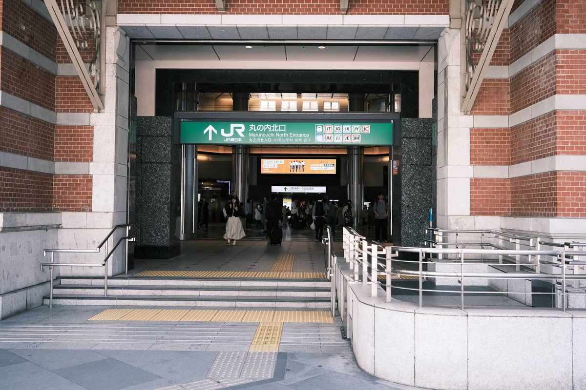Entrance to a JR railway station in Osaka with people walking in, green signage, and brick walls, showing typical city transit access.