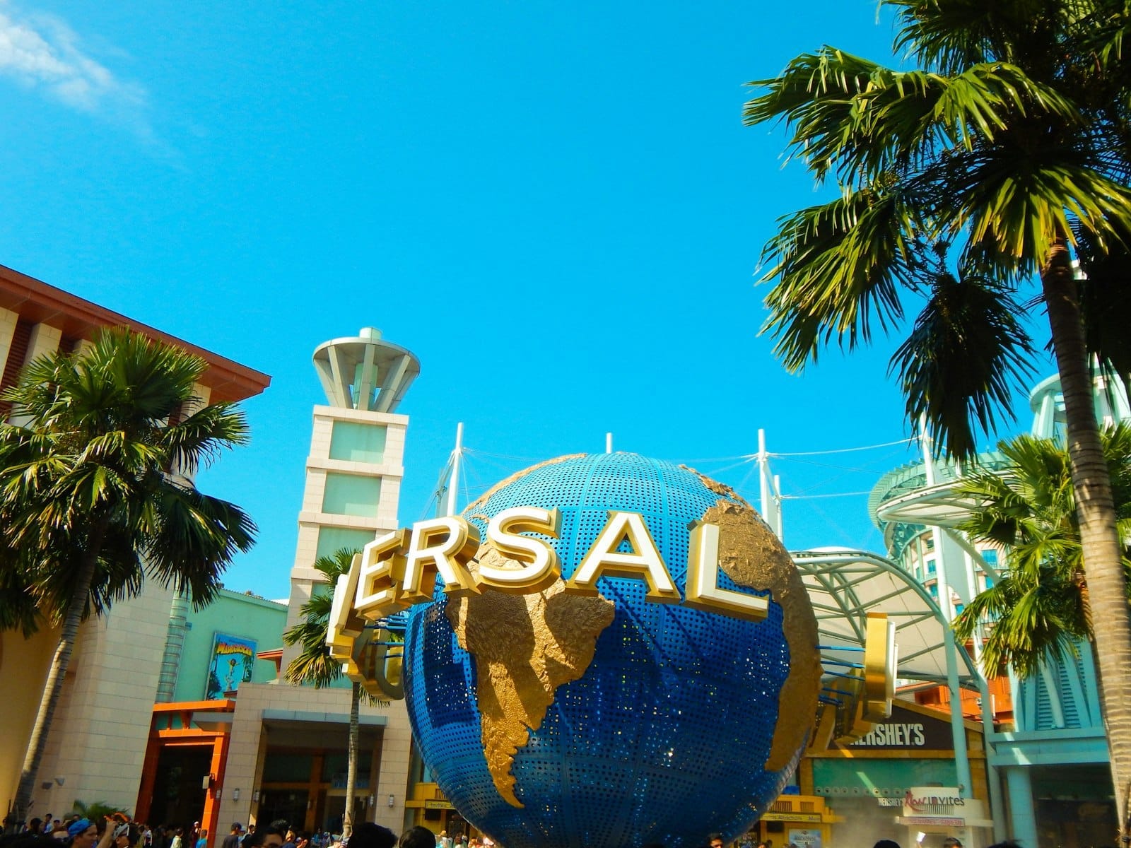 ทางเข้า Universal Studios Singapore มีลูกโลกหมุนอันเป็นสัญลักษณ์ ต้นปาล์ม อาคารสมัยใหม่ และท้องฟ้าสีฟ้าใส