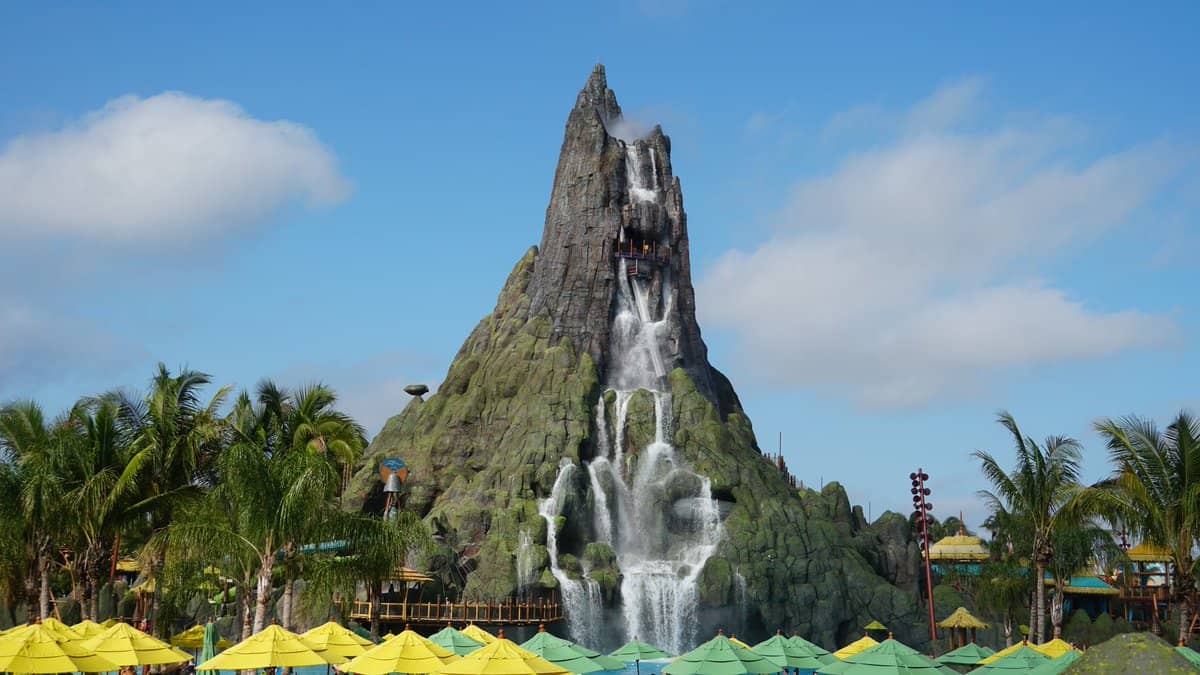 El volcán Krakatau de Universal Volcano Bay con cascadas y palmeras tropicales, visto detrás de coloridas sombrillas bajo un cielo azul.