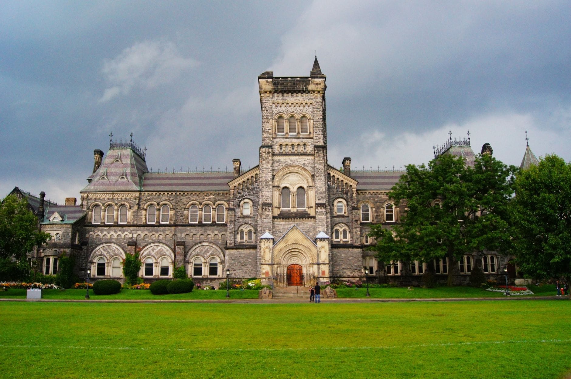 มุมหน้าของ University College ในแคมปัส St. George มหาวิทยาลัยโตรอนโต อาคารหินสไตล์นอร์แมน-โรมานเนสก์ มีหอคอยกลางและสนามหญ้ากว้างใต้ท้องฟ้าเปลี่ยนสี