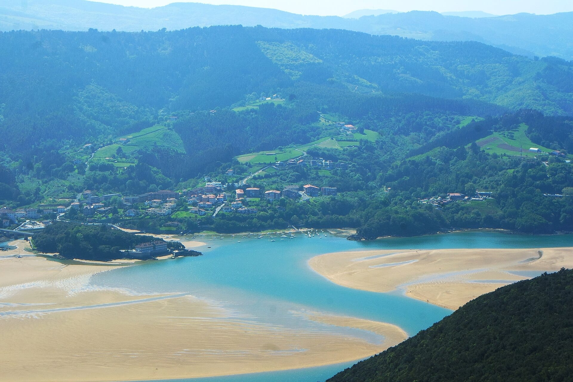 Luftaufnahme des Biosphärenreservats Urdaibai mit dem gewundenen Delta des Flusses Oka, sandigem Ästuar, kleinen Booten und grünen Hügeln mit vereinzelten Häusern.