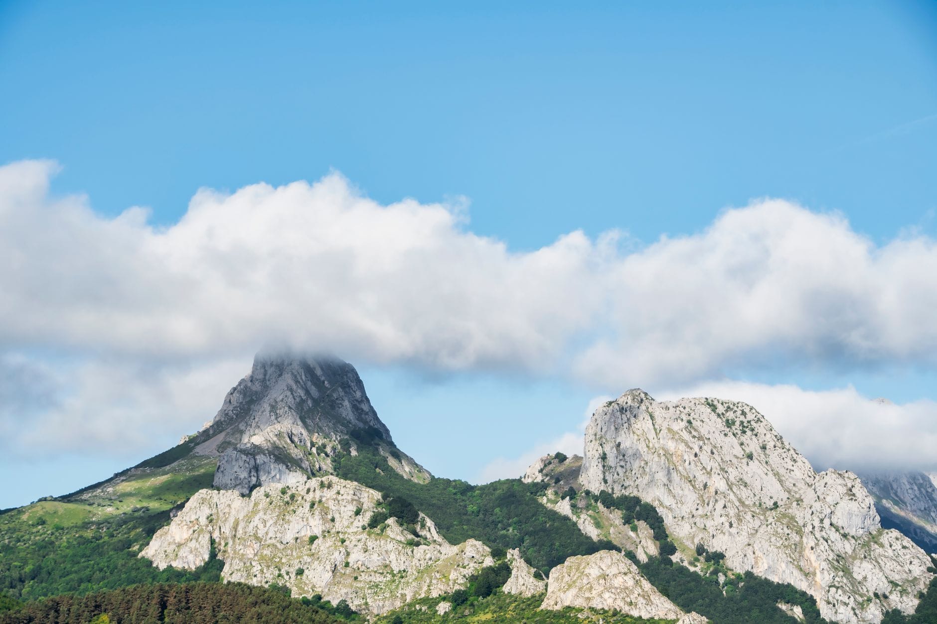 Dramatic limestone peaks of Urkiola Natural Park with lush greenery and a cloud partially covering one summit under a bright blue sky.