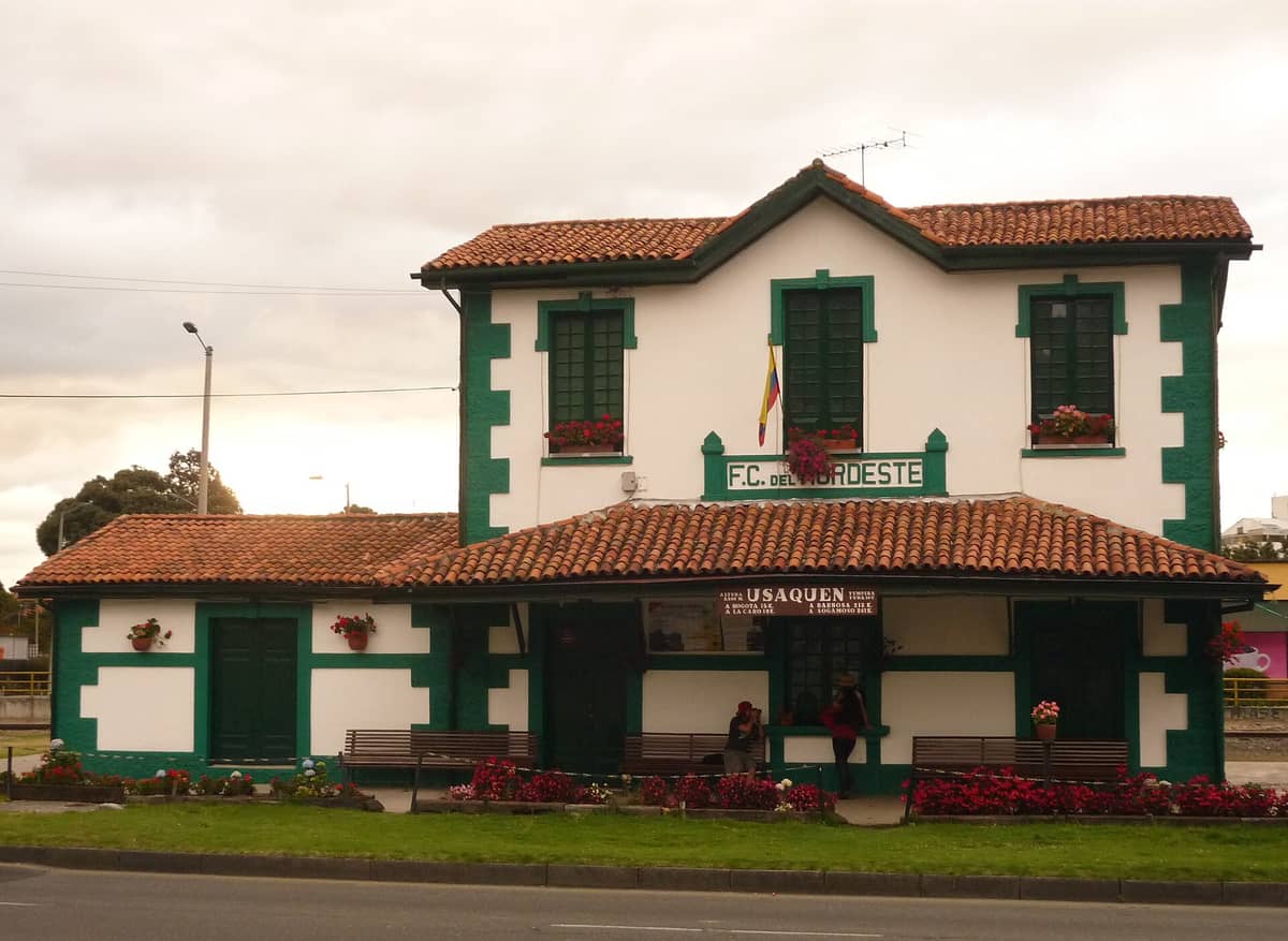 Historic Usaquén train station building with white walls, green trim, and red roof, decorated with flowers and benches, in Bogotá's Usaquén district.