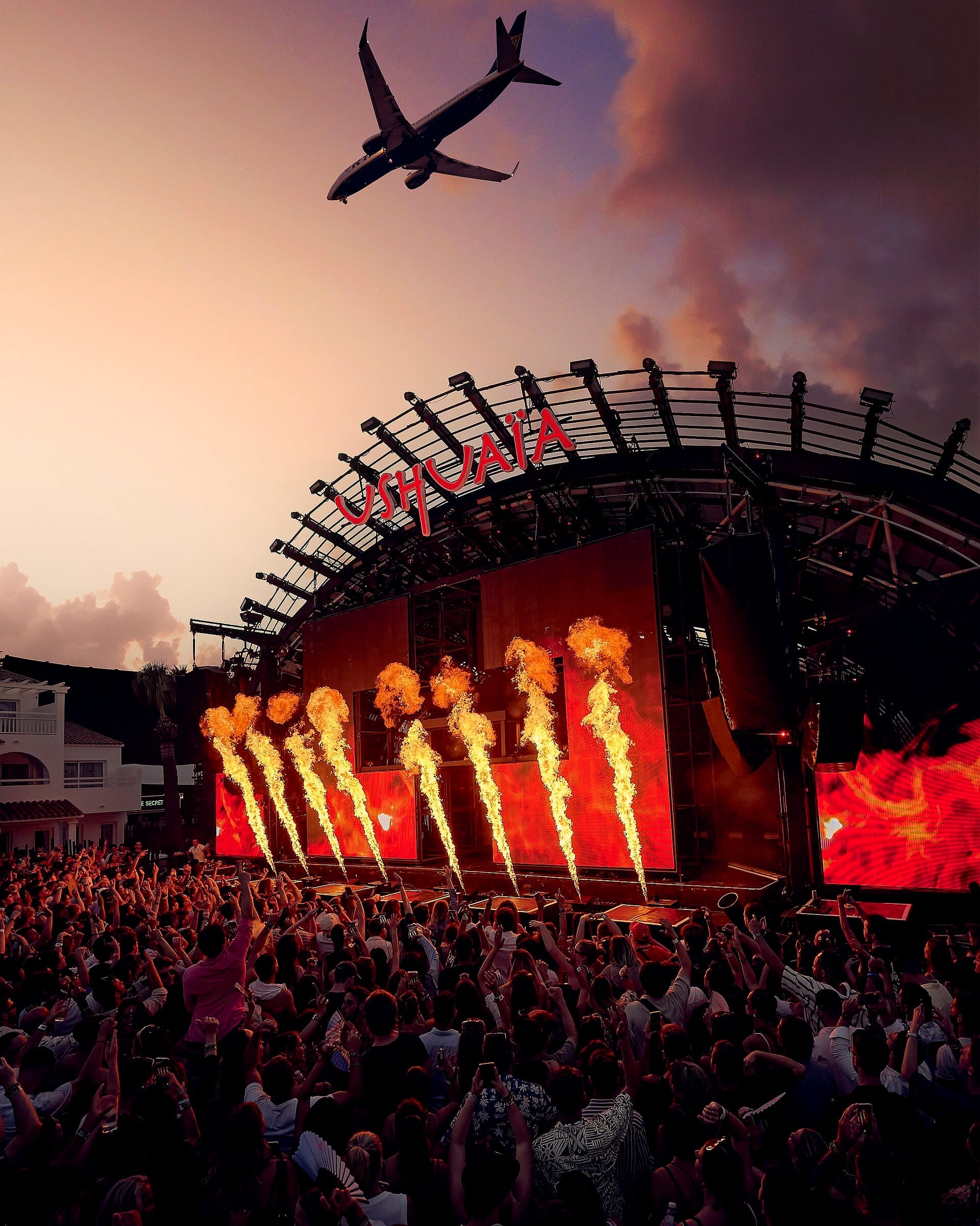 Grande folla all'Ushuaïa Ibiza Beach Hotel che assiste a uno spettacolo sul palco al tramonto con effetti di fuoco e un aereo che vola sopra.