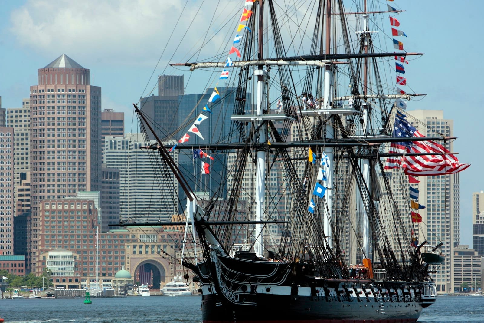 USS Constitution, adornata con variopinte bandiere di segnalazione e una grande bandiera americana, ormeggiata nel porto di Boston con lo skyline della città sullo sfondo.