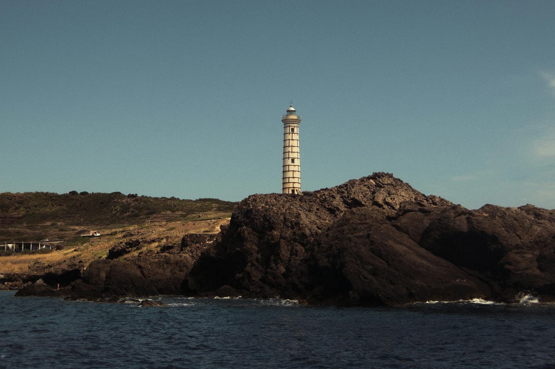 Ein hoher Leuchtturm thront auf dramatischen Vulkanfelsen, umgeben vom tiefblauen Meer unter klarem Himmel – ein Bild, das Usticas maritimen und vulkanischen Charakter perfekt einfängt.