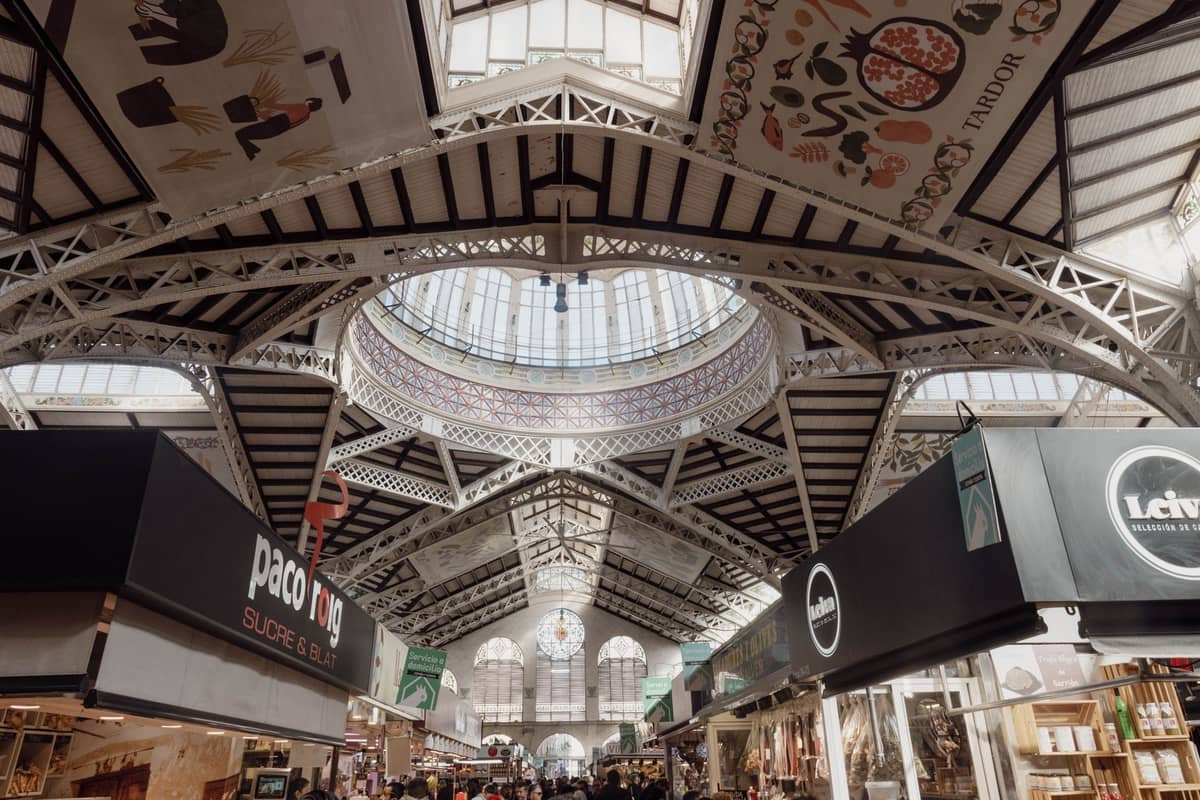Vista interior do Mercado Central de Valencia com ornamentado teto de ferro e vidro, vitrais e detalhes decorativos em azulejos cerâmicos acima das movimentadas bancas do mercado.