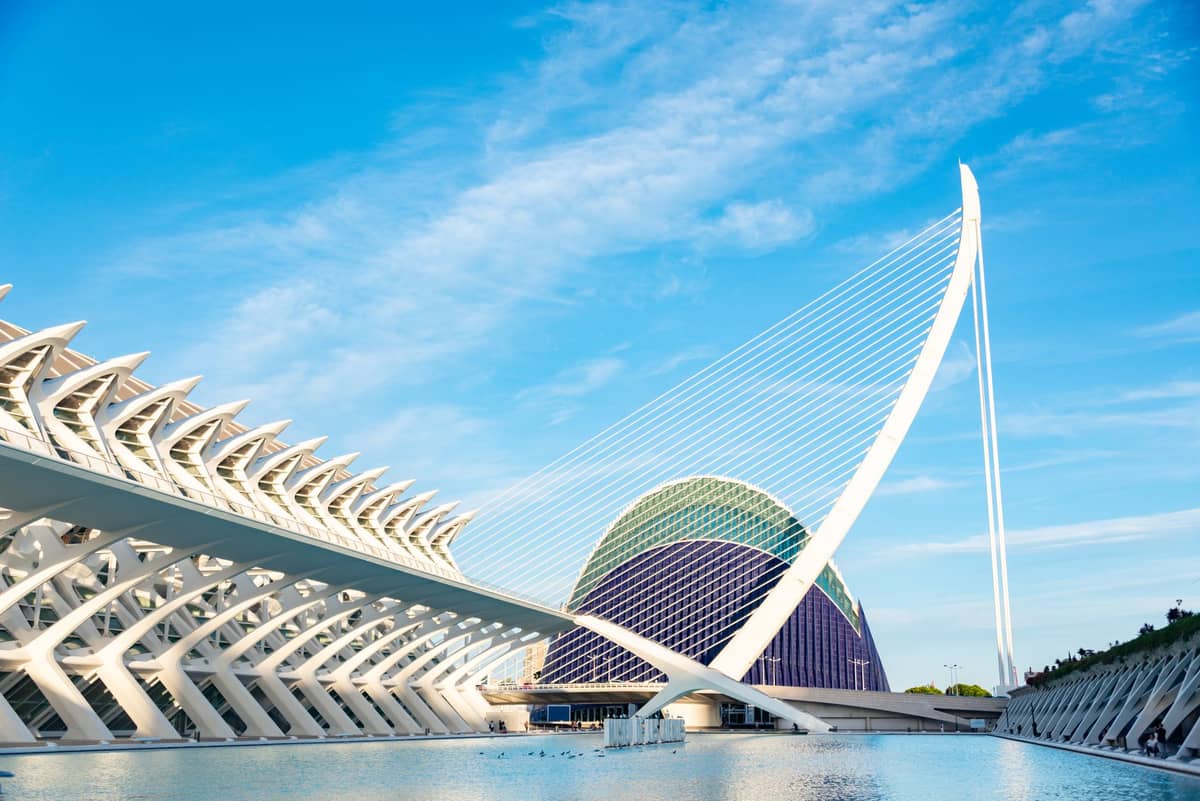 Vista panorâmica da Cidade das Artes e das Ciências em Valência com céu azul, apresentando modernas estruturas esqueléticas brancas e cúpulas de vidro refletidas na água.