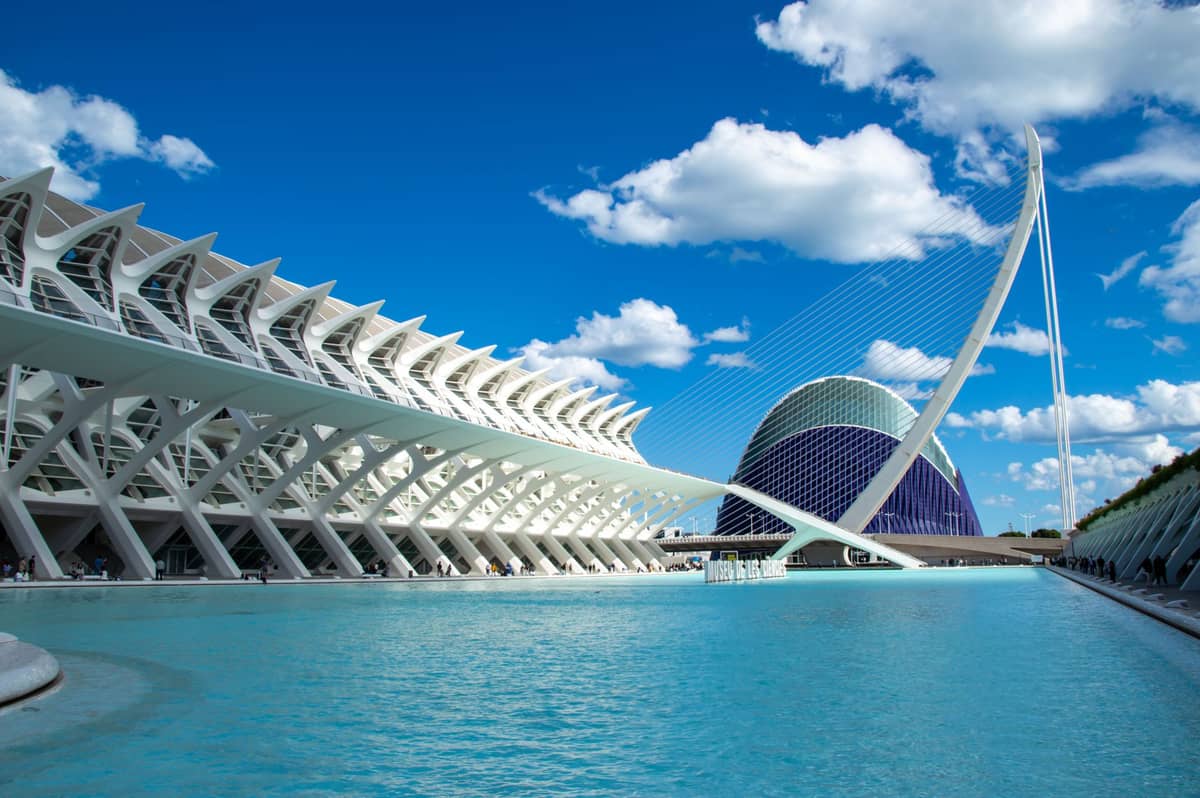Vista panoramica del complesso Città delle Arti e delle Scienze di Valencia: il museo scientifico bianco dalle strutture scheletriche, il suggestivo ponte Assut de l'Or a forma di arpa e la cupola dell'Hemisfèric su uno sfondo di cielo azzurro con nuvole.