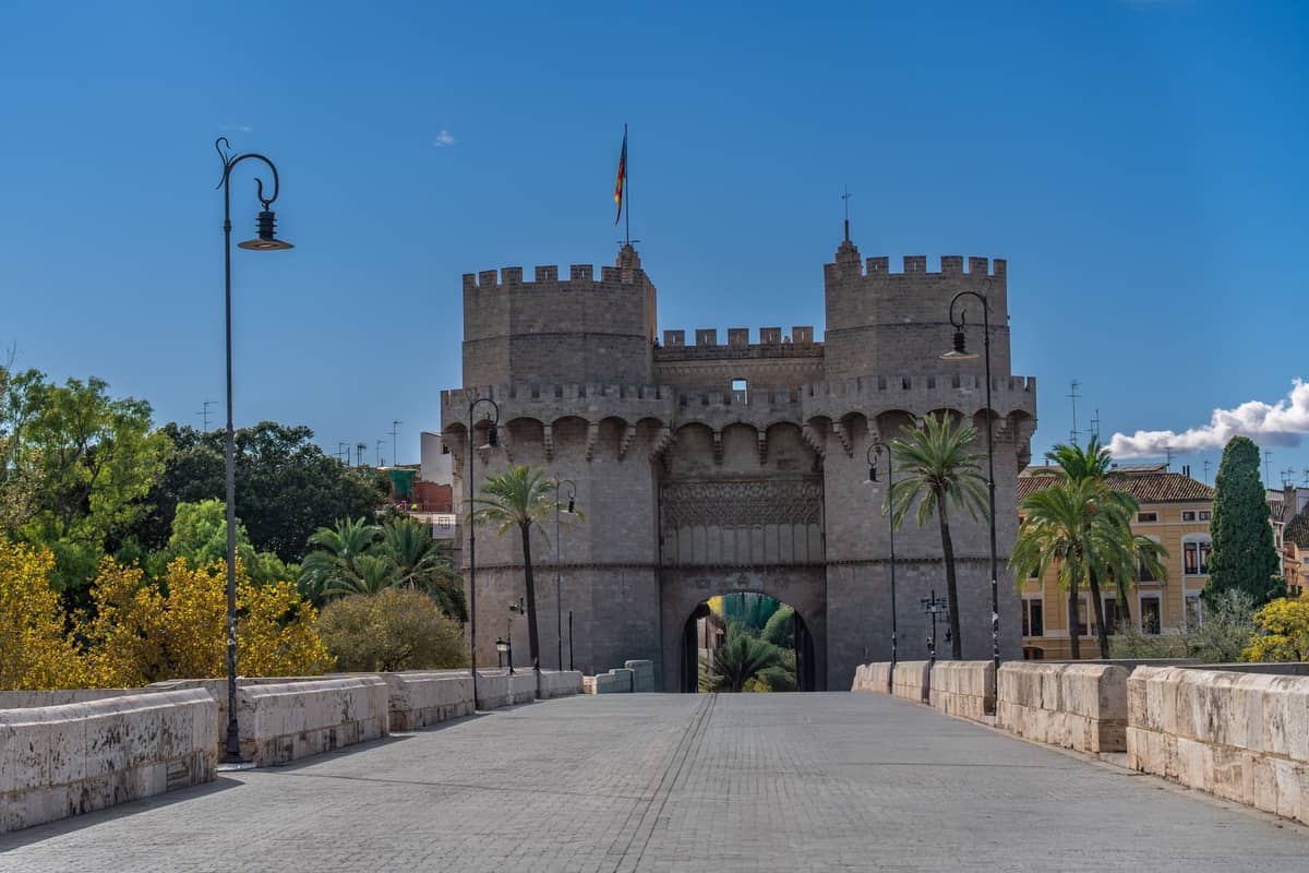 Vista ampla das Torres de Serranos, um portal medieval em Valencia, visto do distrito histórico com árvores ao redor e ponte de pedra.