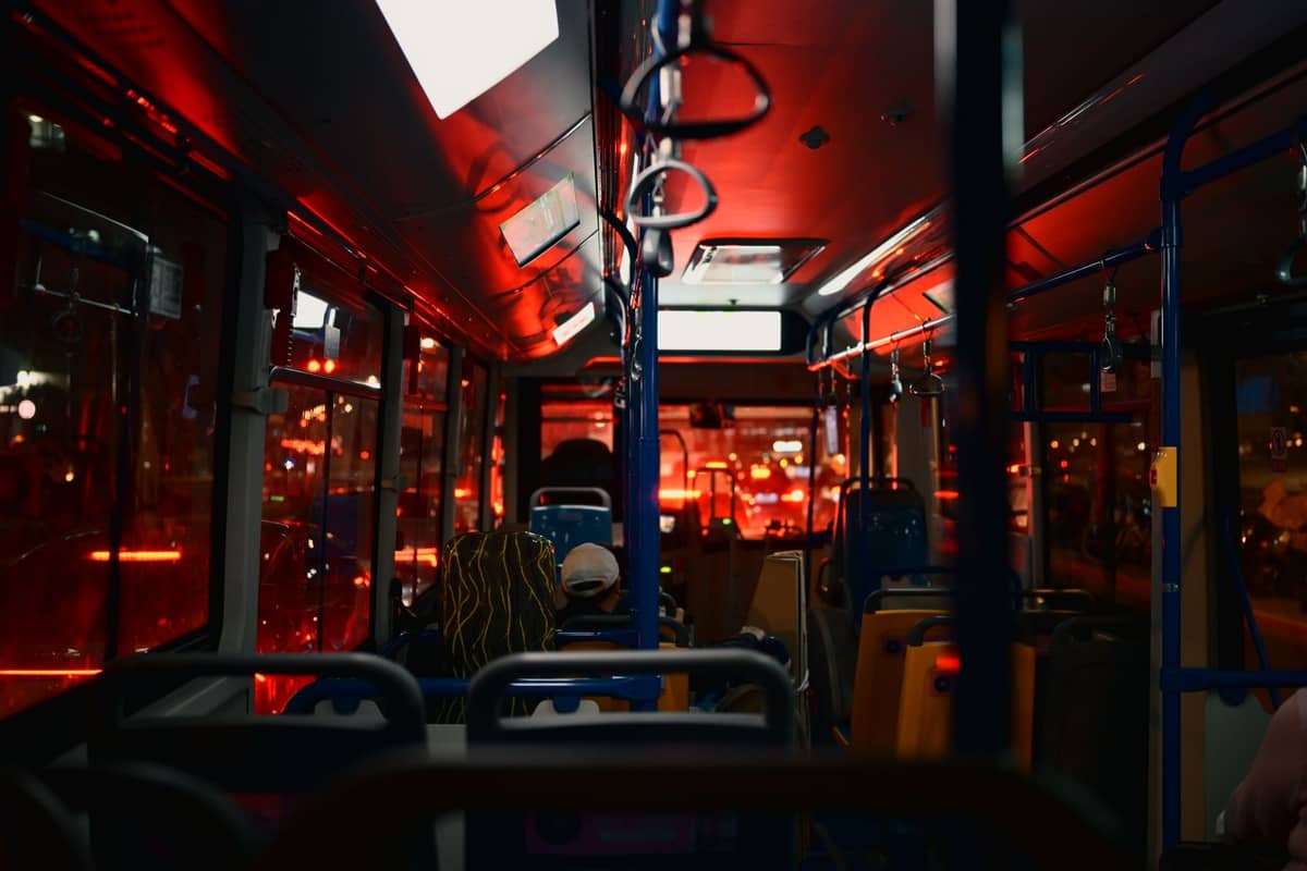Innenraum eines Stadtbusses bei Nacht mit reflektierenden roten Lichtern, Fahrgäste auf ihren Sitzen und Stadtlichter draußen.