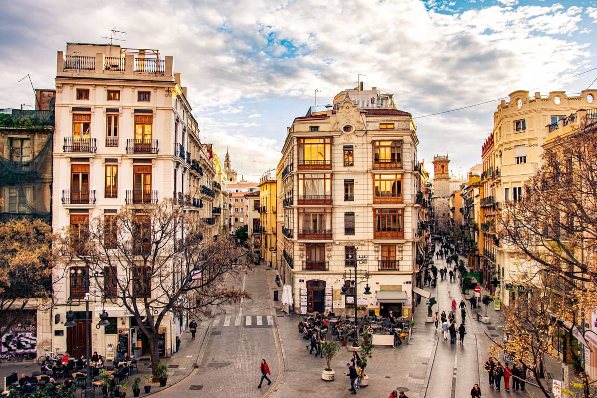Eine lebhafte städtische Straßenszene in Valencia mit Menschen, die sich in Straßencafés versammeln und unter einem teils bewölkten Himmel spazieren.