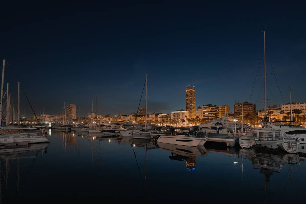 Marina Valencia bei Nacht mit angedockten Yachten und beleuchteten Stadtgebäuden im Hintergrund vor einem dunkelblauem Himmel.