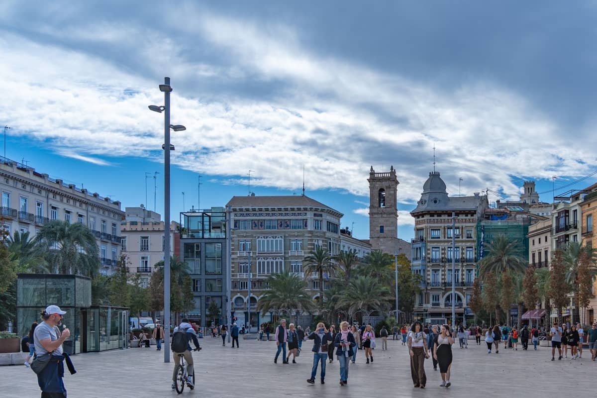Plaza concurrida en Valencia, España, con grupos de personas, palmeras, edificios históricos y un animado ambiente urbano.