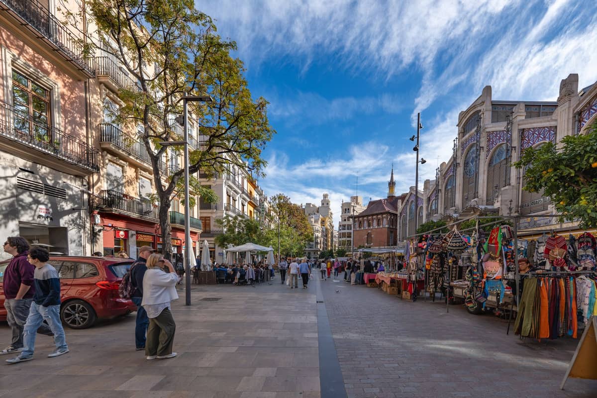Ampia via pedonale dello shopping accanto al Mercado Central di Valencia, con bancarelle all'aperto, l'edificio del mercato e persone che esplorano i negozi sotto un cielo azzurro.