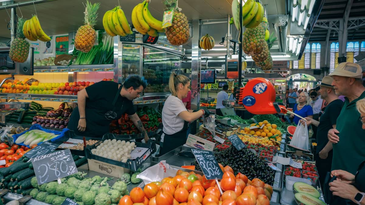 Interior do Mercado Central de Valencia, mostrando bancas vibrantes de frutas e legumes, bananas penduradas, vendedores ocupados e compradores rodeados pela agitação do mercado.