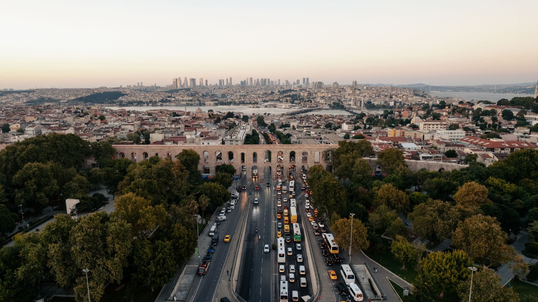Vista aerea dell'Acquedotto di Valente che si estende lungo il Viale Atatürk a Istanbul, circondato da alberi, con lo skyline della città e l'acqua sullo sfondo al tramonto.